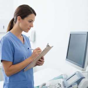 A female patient care technician in blue scrubs intently checks a medical monitor while holding a clipboard and pen.